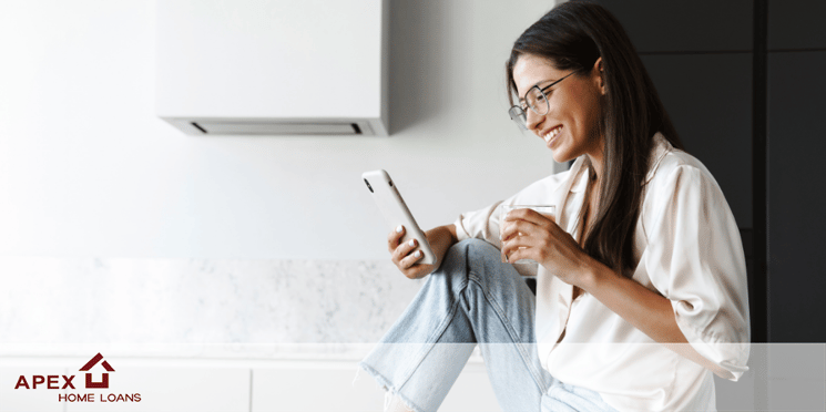 A young woman sitting on her kitchen counter, smiling at her phone as she reads about 2022 home prices.
